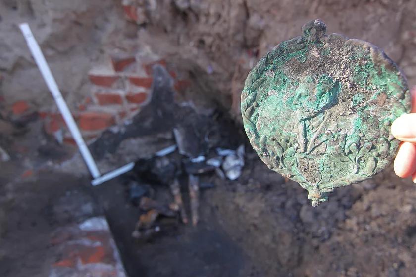 eine Hand hält im Vordergrund eine grün korrodierte Plakette mit dem Gesicht von August Bebel darauf